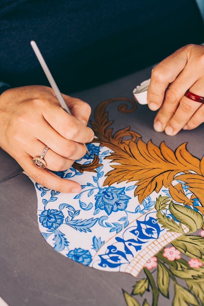 Home Close-up of skilled hands painting intricate floral patterns on pottery, showcasing craftsmanship.